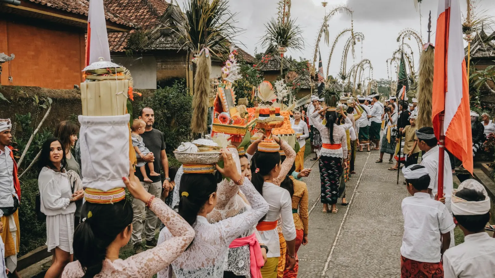A procession of women carrying offerings on their heads along a village street lined with curved bamboo penjor poles