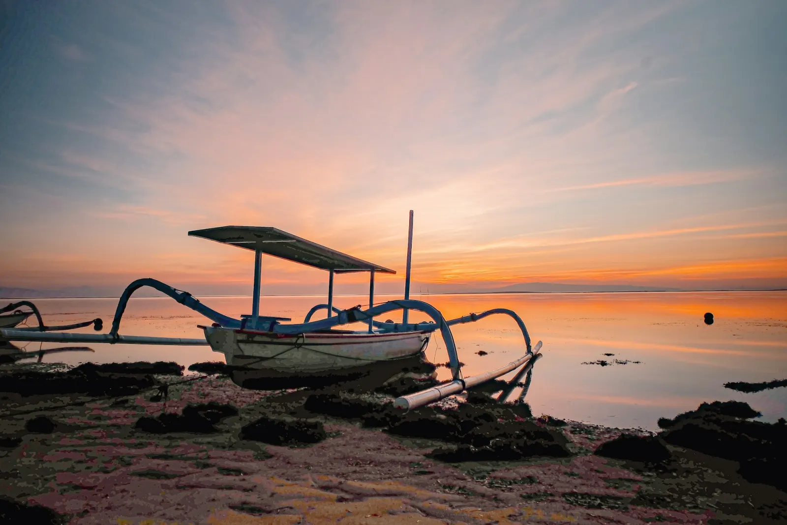 A traditional outrigger fishing boat resting on the rocky shore at sunrise, the sky orange over flat water