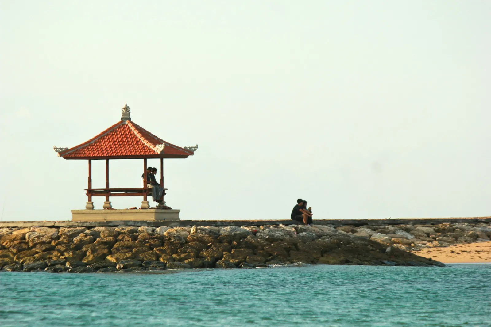 Two people sitting on a stone breakwater beside a red-roofed Balinese pavilion, calm turquoise water in front