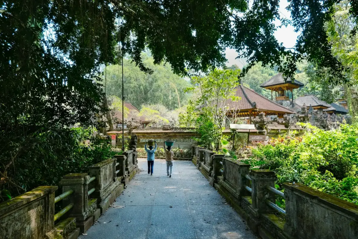 Two people carrying baskets on their heads walk across a stone bridge towards a temple, shaded by tall trees