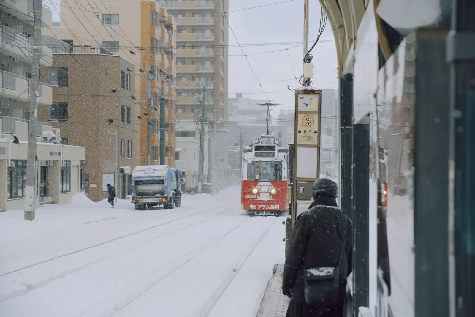 A Sapporo tram pulling into a stop on a snow-covered city street, people waiting under the shelter