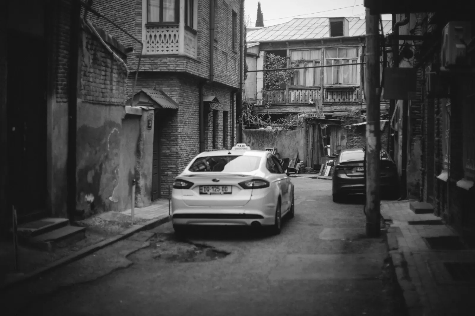 A narrow brick alley in Tbilisi's old town with a white taxi parked under wooden balconies